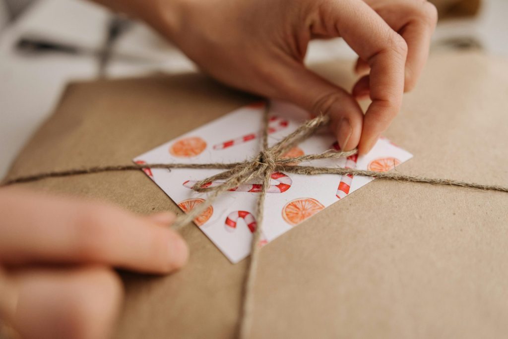 A close-up photo of hands tying a jute rope on a brown paper-wrapped gift with decorated tag.